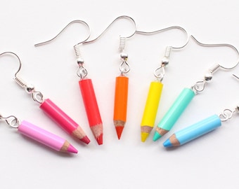 May include: Rainbow colored pencil earrings. The earrings are silver and feature a pink, red, orange, yellow, turquoise, and blue pencil charm.