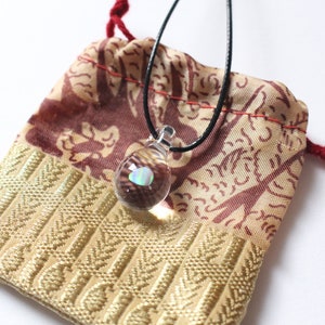 Puede incluir: Un pequeño colgante de vidrio transparente con una piedra de ópalo iridiscente en su interior, colgando de un cordón negro. El colgante está en una pequeña bolsa de cordón rojo y dorado.