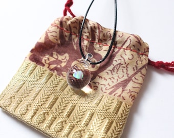 May include: A small, clear glass pendant with an iridescent opal stone inside, hanging from a black cord. The pendant is in a small, red and gold drawstring bag.