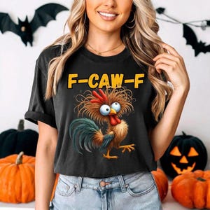 Puede incluir: Camiseta negra con un dibujo de un gallo de dibujos animados y el texto "F-CAW-F" en amarillo. El gallo tiene ojos azules, una cresta roja y una cola verde y azul. La camiseta la lleva una persona delante de decoraciones de Halloween.
