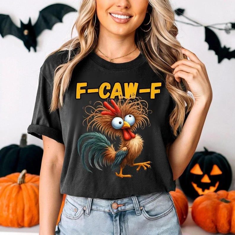 Puede incluir: Camiseta negra con un dibujo de un gallo de dibujos animados y el texto "F-CAW-F" en amarillo. El gallo tiene ojos azules, una cresta roja y una cola verde y azul. La camiseta la lleva una persona delante de decoraciones de Halloween.