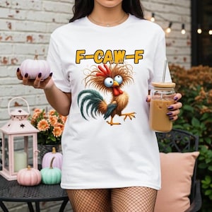 Puede incluir: Camiseta blanca con un dibujo de gallo de dibujos animados y el texto "F-CAW-F" en amarillo. El gallo tiene plumas rojas, marrones y azules. La camiseta es de estilo casual.