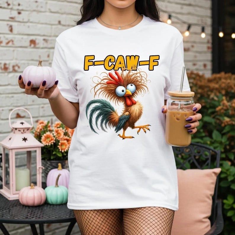 Puede incluir: Camiseta blanca con un dibujo de gallo de dibujos animados y el texto "F-CAW-F" en amarillo. El gallo tiene plumas rojas, marrones y azules. La camiseta es de estilo casual.