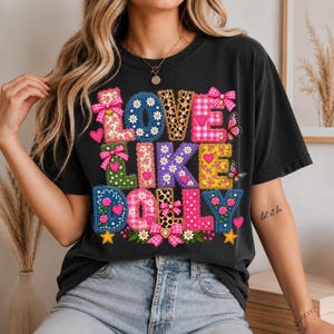 May include: A black t-shirt featuring a colorful graphic print with the text LOVE LIKE DOLLY. The letters are decorated with various patterns including floral, leopard print, gingham, and polka dots, accented with pink bows, hearts, and butterflies.