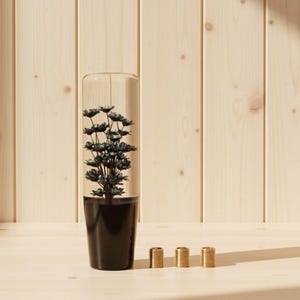 以下が含まれることがあります： 黒い花のアレンジメントが、黒いベースの透明なガラスの花瓶に入っています。3つの小さな金色の円筒形の物体が花瓶の前に並んでいます。背景は明るい色の木の壁とテーブルです。