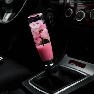 Könnte beinhalten: Ein individueller Autoschaltknauf mit einem transparenten Harzkörper, gefüllt mit rosa Kirschblüten. Die Basis des Knaufs ist einfarbig rosa. Der Schaltknauf ist in einem schwarzen Autointerieur installiert.