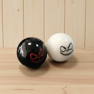 Könnte beinhalten: Zwei kugelförmige Dekorationsgegenstände. Einer ist schwarz mit einem roten Gesichtsdesign, der andere ist weiß mit einem schwarzen Gesichtsdesign. Die Objekte sind auf einer Holzoberfläche platziert und wahrscheinlich zum Thema Halloween.