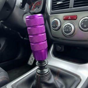 Pode incluir: Um botão de mudança de marcha roxo para carro com um design cilíndrico texturizado. O botão está instalado no interior de um veículo, com o painel e outros controles visíveis ao fundo. Um acessório de carro elegante.