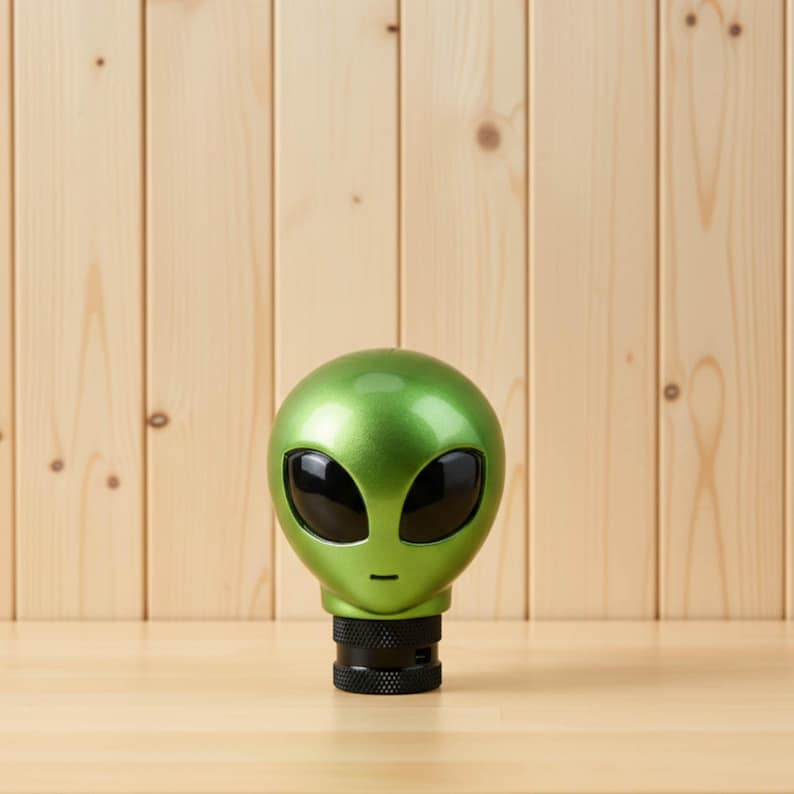 Green alien head shift knob installed on a manual transmission car, close-up on the glossy resin finish with black eyes and textured base adapter; custom shift knob for 5/6-speed gear shifter, JDM style accent for interior upgrades, ideal for sim racing, iRacing and gaming simulator cockpits; ergonomic weighted shifter knob for precise shifts; universal fit with adapters for common threads; unique sci-fi gift for car enthusiasts and tuners.