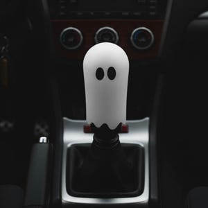 White ghost weighted shift knob on a manual transmission gear shifter in a dark car interior, cute Halloween JDM shift knob and custom gear shifter knob for 6 speed cars, manual shift knob upgrade for drift builds, stance cars and hot rods, unique gear knob for sim racing cockpit, iRacing rig or gaming simulator setup, funny spooky car accessory for modified JDM interiors and tuners.