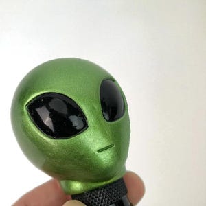 Handheld close-up of green alien head shift knob highlighting pearlescent resin texture and glossy black eyes; custom shift knob for manual transmission gear shifters, designed for 6-speed and 5-speed cars; durable, weighted feel for better control; ideal interior upgrade for JDM style projects, street cars and drift builds; also popular for sim racing, iRacing and gaming simulator use; universal adapters make installation quick and easy.
