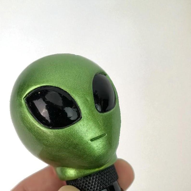 Handheld close-up of green alien head shift knob highlighting pearlescent resin texture and glossy black eyes; custom shift knob for manual transmission gear shifters, designed for 6-speed and 5-speed cars; durable, weighted feel for better control; ideal interior upgrade for JDM style projects, street cars and drift builds; also popular for sim racing, iRacing and gaming simulator use; universal adapters make installation quick and easy.
