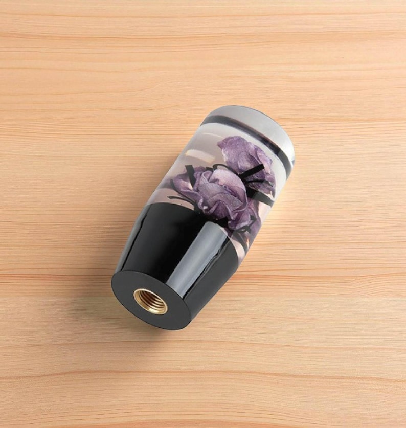 Close up of purple rose resin shift knob lying on its side showing threaded metal insert, custom shift knob for manual transmission gear shifter, clear resin body with embedded purple flowers, weighted gear knob compatible with 6 speed cars, ideal for sim racing, iRacing cockpit and gaming simulator shifter upgrades.