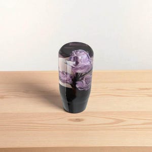 Front view of purple roses resin shift knob 4 inch tall on wooden table, clear acrylic flower shift knob with three detailed violet roses inside, weighted shift knob design for smooth gear changes, universal manual transmission shifter knob for JDM cars, hot rods, custom builds and sim racing gear shifter setups.