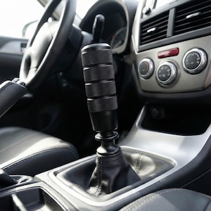 May include: A black, textured shift knob installed in a car. The shift knob has a cylindrical design with a series of grooved rings. The interior of the car features a black leather gear shift boot, a steering wheel, and a silver dashboard.