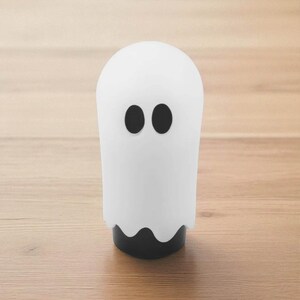 Standalone white ghost weighted shift knob standing on a wooden surface, minimalist Halloween gear knob design with black oval eyes and wavy base, JDM inspired custom shift knob for manual transmission cars and 6 speed gear shifters, funny interior car accessory for hot rod and rat rod builds, stance projects and drift cars, also ideal as a sim racing or gaming simulator ghost shifter knob for unique spooky cockpit style.