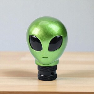 Front view of metallic green alien head shifter knob on a wooden surface, showing smooth resin contours and large black eyes; custom gear shift knob compatible with manual transmission vehicles (5/6-speed) and threaded automatic levers; perfect for JDM builds, track toys and daily drivers; premium resin construction with comfortable grip for accurate gear changes; great accessory for sim racing rigs, iRacing setups and gaming simulator cockpits.