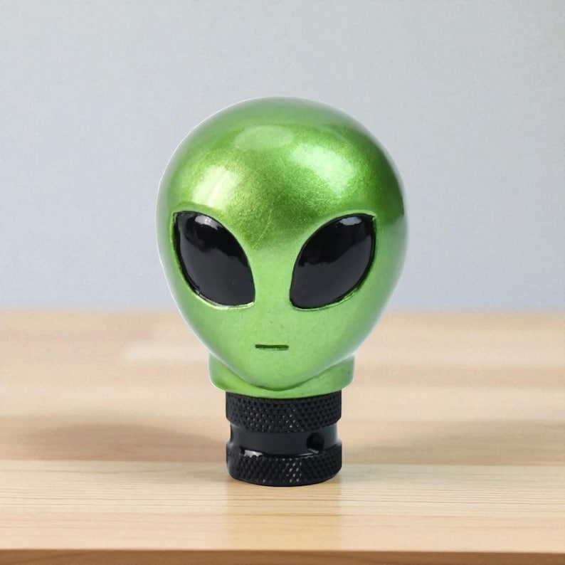Front view of metallic green alien head shifter knob on a wooden surface, showing smooth resin contours and large black eyes; custom gear shift knob compatible with manual transmission vehicles (5/6-speed) and threaded automatic levers; perfect for JDM builds, track toys and daily drivers; premium resin construction with comfortable grip for accurate gear changes; great accessory for sim racing rigs, iRacing setups and gaming simulator cockpits.