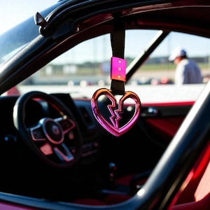 May include: A pink and orange heart-shaped car handle with a broken heart design. The handle is suspended from a black strap and mounted inside a red car. The steering wheel and dashboard are visible.