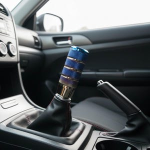 以下が含まれることがあります： 青とシルバーの車のギアシフトノブ、テクスチャードグリップ付き。 シフトノブは車の内装に設置されており、黒革のシフトブーツと黒いハンドブレーキレバーが見えます。