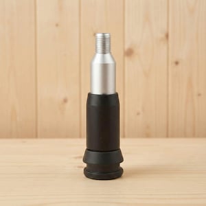 Könnte beinhalten: Ein silberner und schwarzer Flaschenverschluss. Der obere Teil ist silberfarben mit einem Gewinde, während der mittlere Teil schwarz ist. Die Basis des Stöpsels hat ein ausgestelltes, abgestuftes Design. Der Hintergrund ist helles Holz.