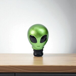 Studio shot of green alien head gear shifter knob standing upright, matte background; resin shift knob with ergonomic shape and textured collar, compatible with most manual shift knob threads and some threaded automatics; standout JDM-inspired accessory for interior customization; enhances grip and shifting feel; great for sim racing rigs, iRacing and gaming simulator setups; creative gift for car lovers, tuners and sci-fi fans.
