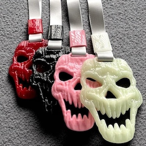 May include: Four skull-shaped pendants in red, black, pink, and off-white, each with a matching ribbon loop. The skulls have detailed features and open mouths, and are suspended from white ribbons.