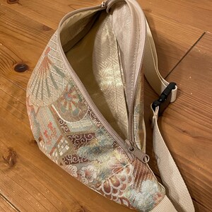 May include: A beige fanny pack with a floral and geometric pattern in gold, brown, and red. It features a zip closure and an adjustable beige strap with a black buckle. The interior lining is a solid beige colour.