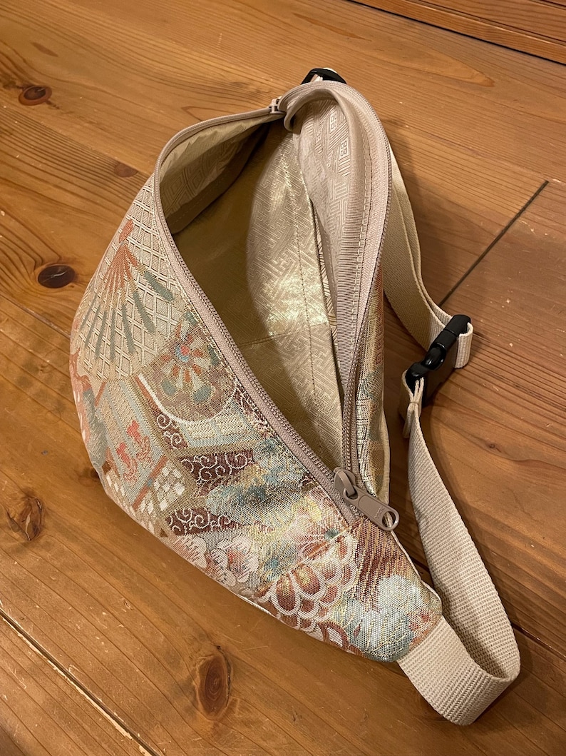 May include: A beige fanny pack with a floral and geometric pattern in gold, brown, and red. It features a zip closure and an adjustable beige strap with a black buckle. The interior lining is a solid beige colour.
