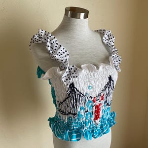 May include: A white crop top with a blue and white polka dot ruffle trim. The top has a white background with a blue and white pattern of a bridge and a red and white boat.