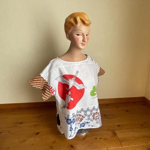 May include: A white short-sleeved top featuring a red circular sun motif, a crane, and a turtle. The sleeves are brown with white polka dots. The lower section displays various blue and brown geometric and floral patterns on a white background.