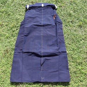 May include: A navy blue hakama, a traditional Japanese garment, lies on green grass. The hakama is rectangular with vertical pleats and a white band at the top. A small, embroidered design in orange is visible on the upper right.