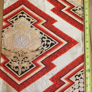 May include: A gold and red fabric with a geometric pattern. The fabric has a black border and a floral design in the center. The fabric is approximately 15 inches wide.