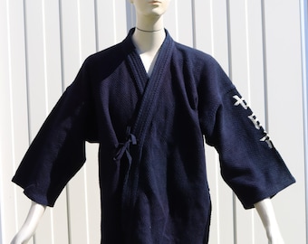 Op de afbeelding: Een donker marineblauw kendo gi-jasje met een getextureerd sashiko-weefsel, een overslagsluiting met stoffen linten en witte Japanse kanji-tekens geborduurd op de linkermouw, gepresenteerd op een etalagepop.