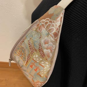 May include: A patterned fanny pack with a cream-coloured strap. The bag features a floral design in shades of brown, orange, and blue. The zipper is silver. The bag is worn across the body.