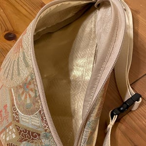 May include: A beige patterned fanny pack with a zip closure. The bag has a cream-coloured strap with a black buckle. The interior lining is a light gold colour. The bag is made of a patterned fabric with floral and geometric designs.