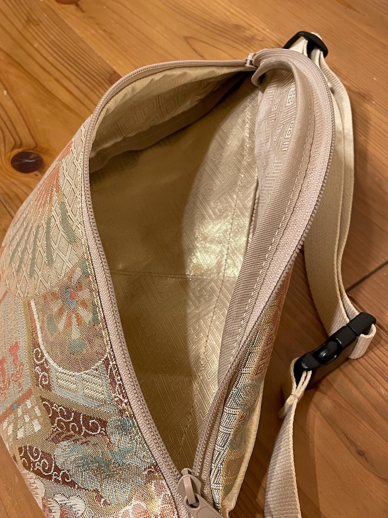 May include: A beige patterned fanny pack with a zip closure. The bag has a cream-coloured strap with a black buckle. The interior lining is a light gold colour. The bag is made of a patterned fabric with floral and geometric designs.