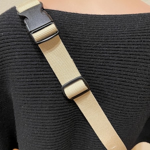 May include: A tan crossbody bag strap with a black plastic buckle and adjuster. The strap is made of woven material and is attached to a black sweater. The strap is approximately 100 cm long.