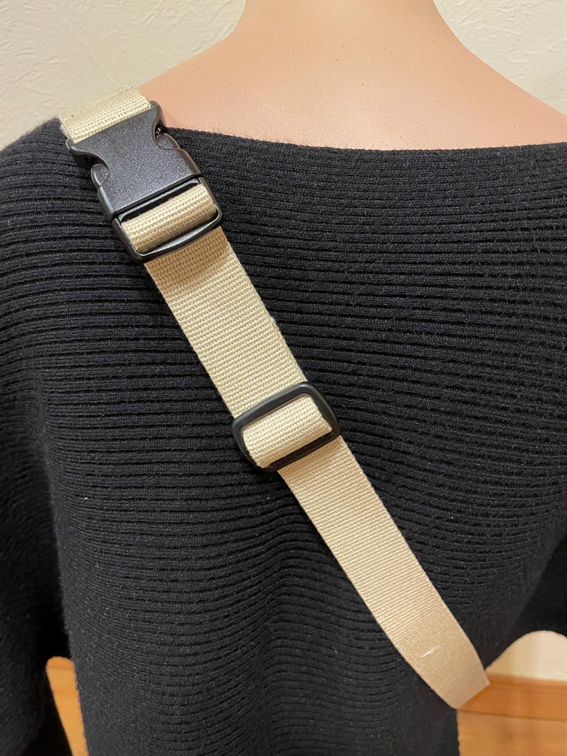 May include: A tan crossbody bag strap with a black plastic buckle and adjuster. The strap is made of woven material and is attached to a black sweater. The strap is approximately 100 cm long.