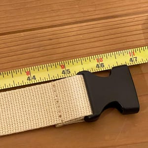 May include: A beige nylon belt with a black plastic buckle. The belt is measured with a yellow tape measure showing 112 to 120 cm. The belt is on a wooden surface.