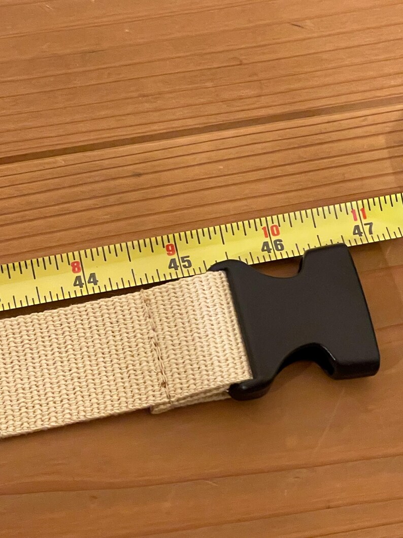 May include: A beige nylon belt with a black plastic buckle. The belt is measured with a yellow tape measure showing 112 to 120 cm. The belt is on a wooden surface.