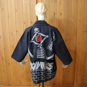 May include: A dark navy blue short-sleeved jacket featuring a white graphic print of a brick wall, a barrel, and a diamond-shaped frame with red Japanese kanji characters. The garment has a wide collar and a black waist tie, displayed on a mannequin.