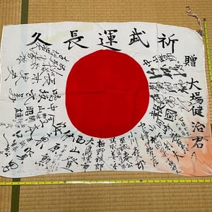 May include: A white Japanese flag with a large red circle in the centre, covered in black ink calligraphy signatures. A yellow measuring tape is placed along the bottom and right edges, showing the flag is approximately 91 cm wide and 71 cm tall.