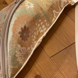May include: Close-up of a beige and floral patterned bag with a zip closure. The bag features a cream-coloured strap and a detailed floral design in shades of orange, green, and beige. The bag is on a wooden surface.