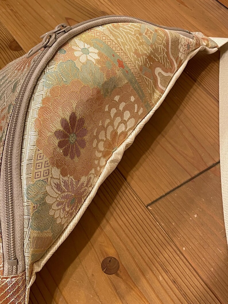 May include: Close-up of a beige and floral patterned bag with a zip closure. The bag features a cream-coloured strap and a detailed floral design in shades of orange, green, and beige. The bag is on a wooden surface.