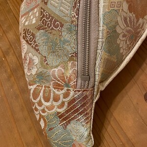 May include: Close-up of a patterned fanny pack with a beige zip and strap. The fabric features a floral design in shades of brown, beige, and teal. The zip is a neutral beige colour. The strap is a solid beige.