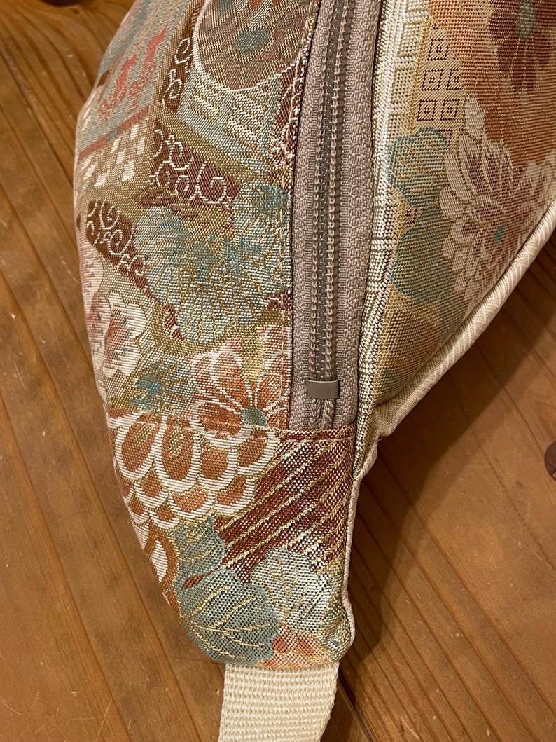 May include: Close-up of a patterned fanny pack with a beige zip and strap. The fabric features a floral design in shades of brown, beige, and teal. The zip is a neutral beige colour. The strap is a solid beige.