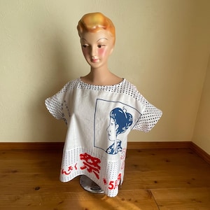May include: A white short-sleeved top with black polka dot patterns on the shoulders and sides. The front features a blue graphic illustration of a person in profile within a square frame, red Japanese kanji text, and additional black text.
