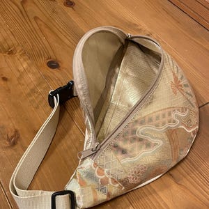 May include: A beige fanny pack with a decorative floral pattern in gold, brown, and orange. The bag features a zippered main compartment, an adjustable beige strap, and a black buckle. The bag is open, showing a gold interior.