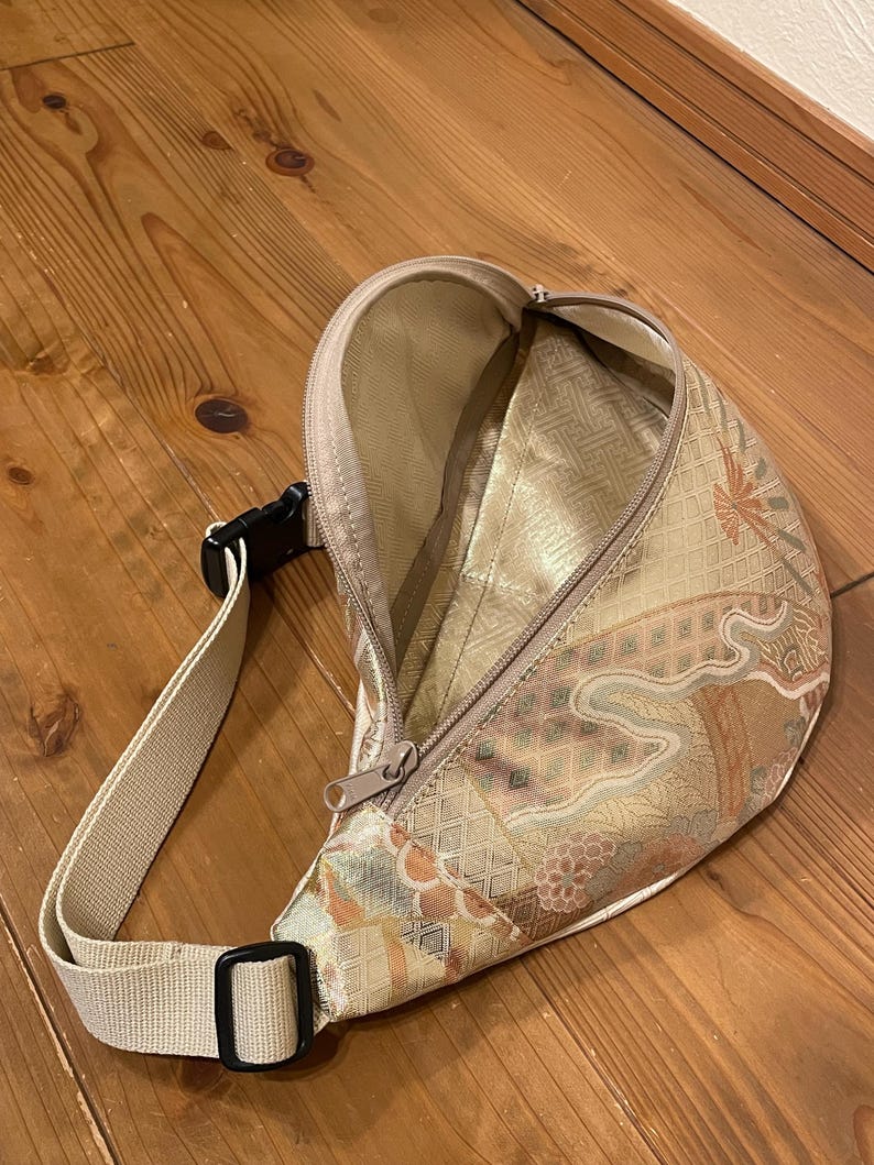 May include: A beige fanny pack with a decorative floral pattern in gold, brown, and orange. The bag features a zippered main compartment, an adjustable beige strap, and a black buckle. The bag is open, showing a gold interior.